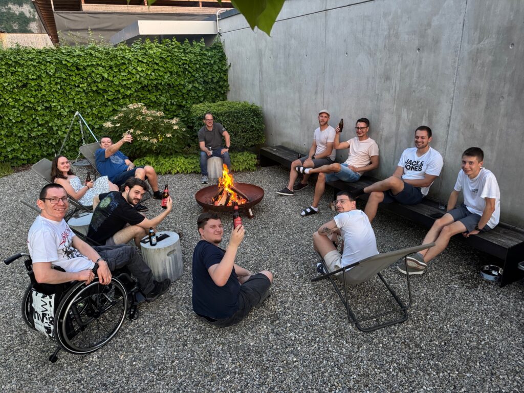 Mitarbeitende von MIA Systems sitzen am Abend gemeinsam um ein Lagerfeuer im Firmenhof, mit Getränken in der Hand und entspannter Stimmung.