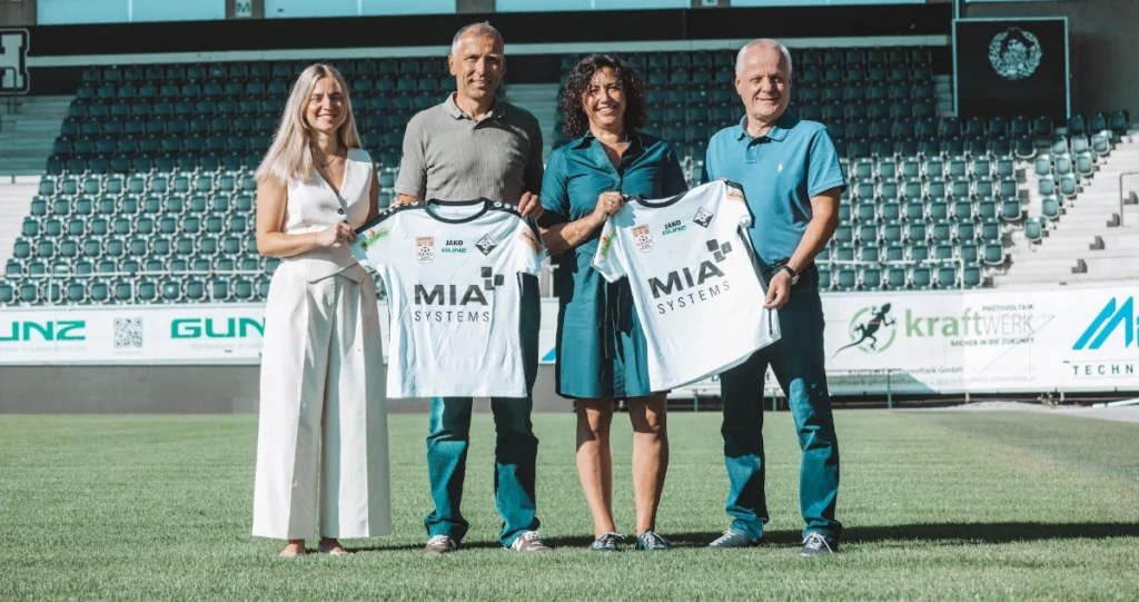 f.l.t.r. Lisa Metzler, sportsleader SCRA, Alexander Fehr, CEO MIA Systems GmbH mit Ulrike Fehr, Werner Gunz, Vicepresident SCRA are standing in the Schnabelholz Stadion an showing two womens soccer shirts with the MIA logo.