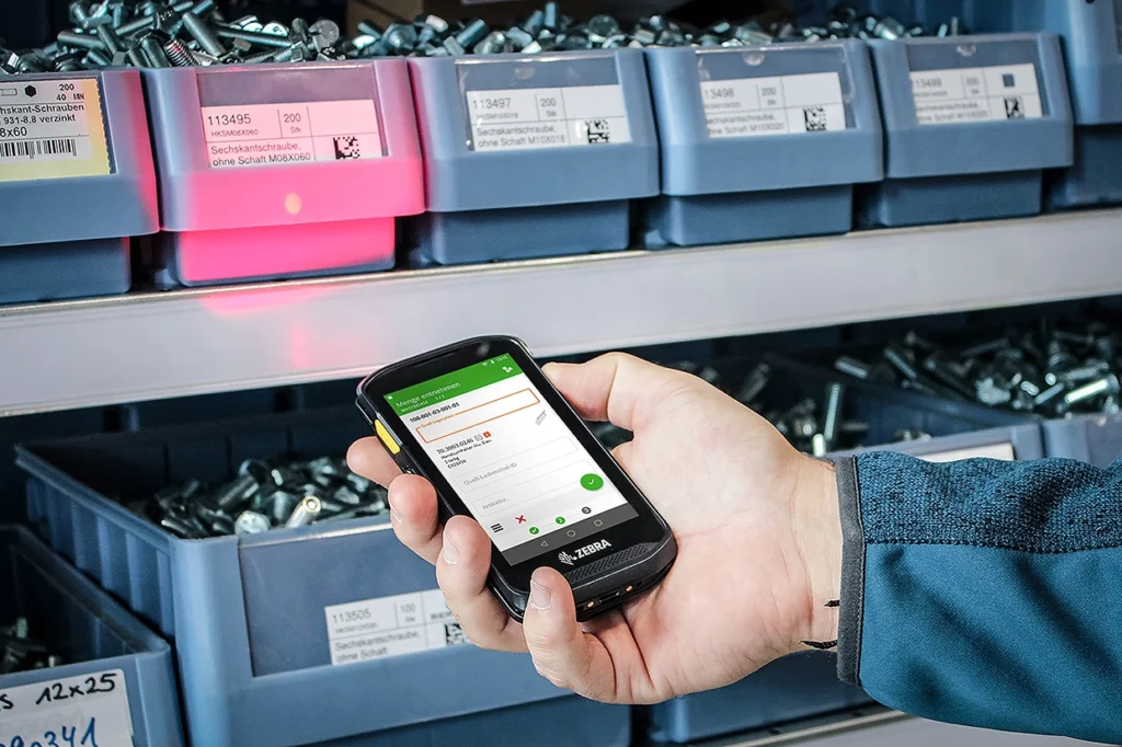 Nahaufnahme eines WMS Mobile in einer Hand, vor einem Lagerregal mit Kleinteilen.