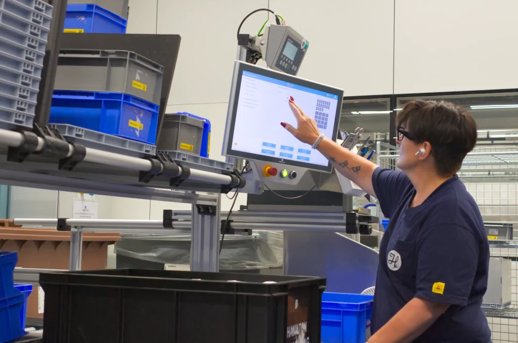 Eine Lagermitarbeiterin bedient an einer Kommissionierungsstation von Servus das MIA Touch. Diese teilweise Lagerautomation beruht auf dem Prinzip Ware zur Person.